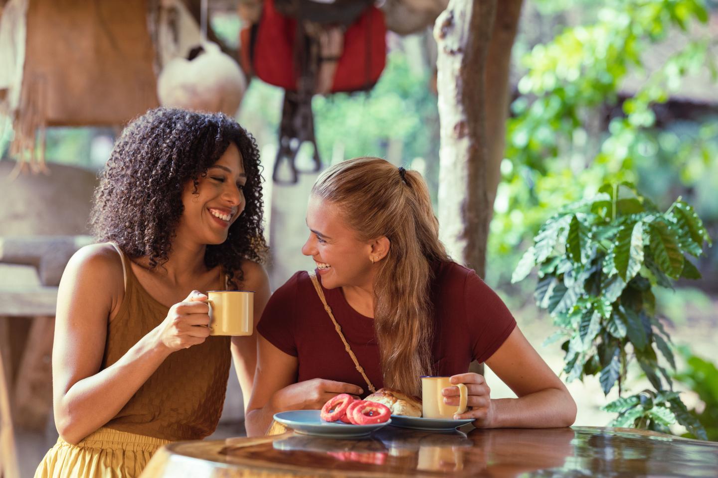 Dos mujeres sonríen mientras toman café y pasteles en una mesa al aire libre.