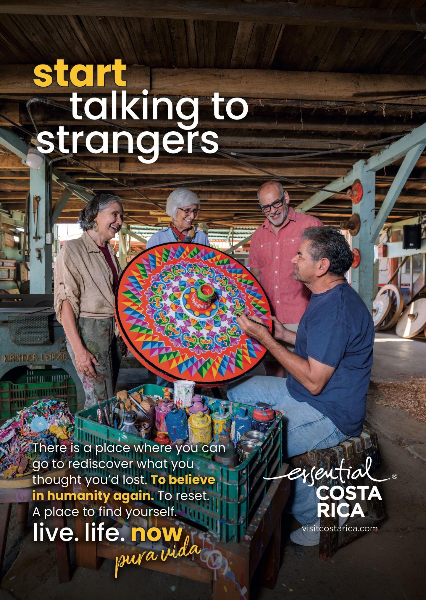 Anuncio en el que aparecen cuatro personas en un taller charlando con un artesano que sostiene una gran pieza artesanal circular pintada con colores vivos. Título: «Empieza a hablar con desconocidos». Marca: «esencial Costa Rica, visitcostarica.com.