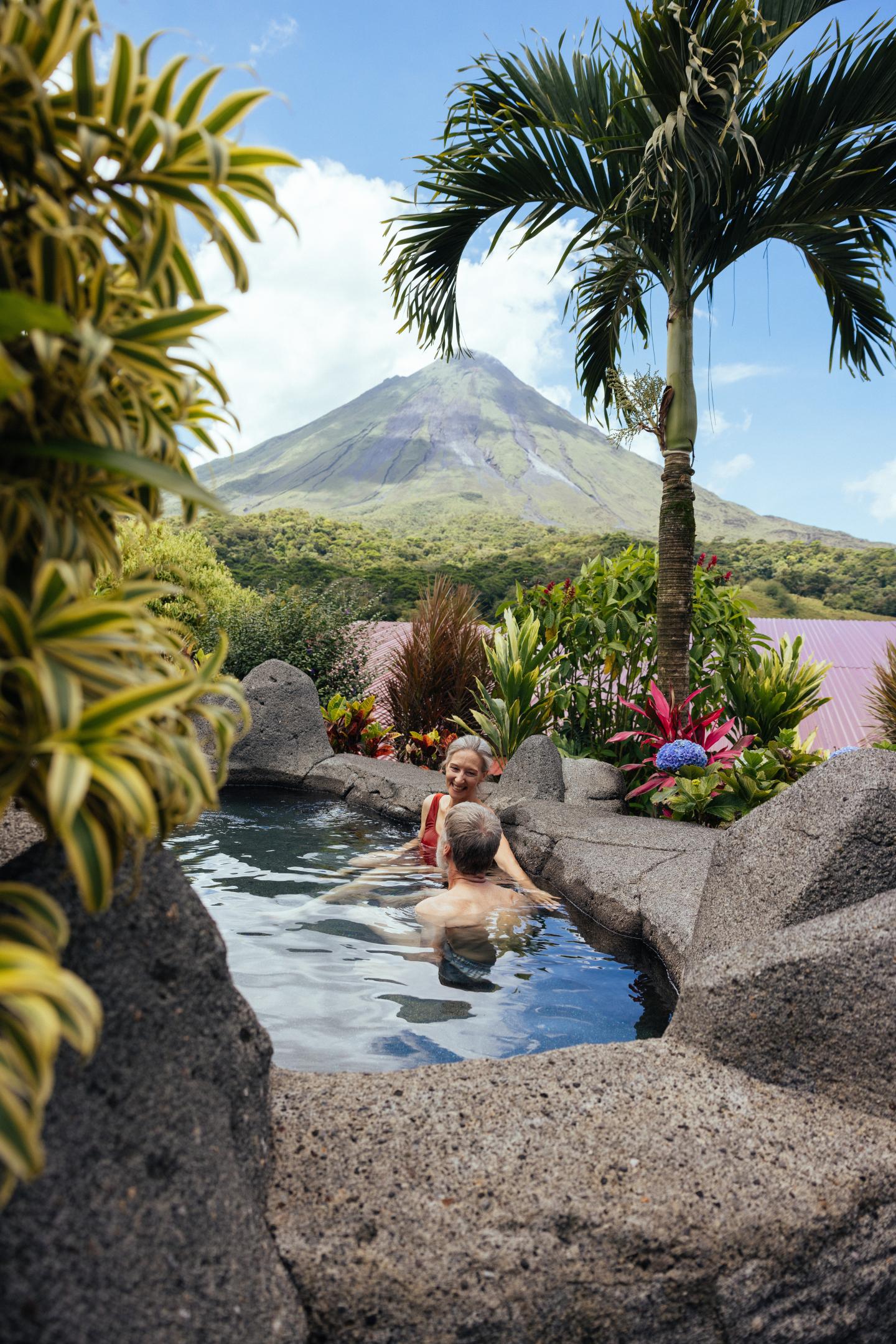 Dos personas se relajan en una piscina termal de piedra rodeada de plantas tropicales, con el volcán Arenal al fondo.