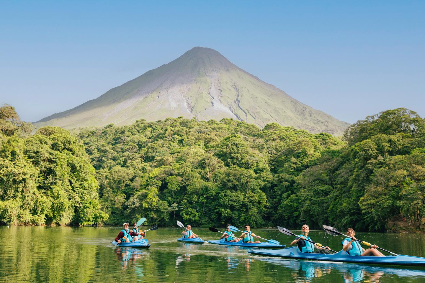 Paseo en kayak en grupo por un lago tranquilo rodeado de selva tropical, con el volcán Arenal asomando al fondo.